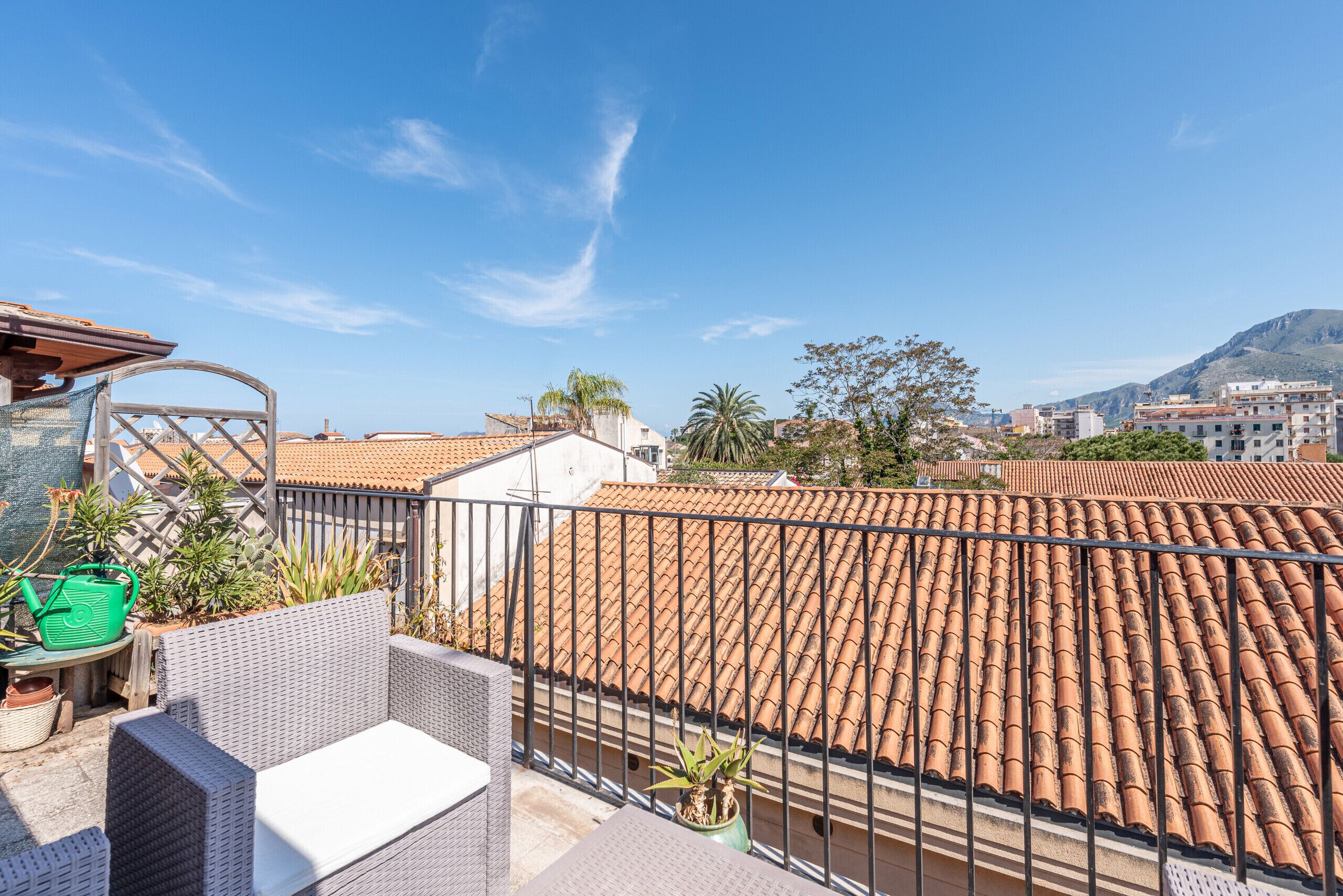 A rooftop terrace with a view of terracotta-tiled buildings, a wicker sofa, and potted plants.