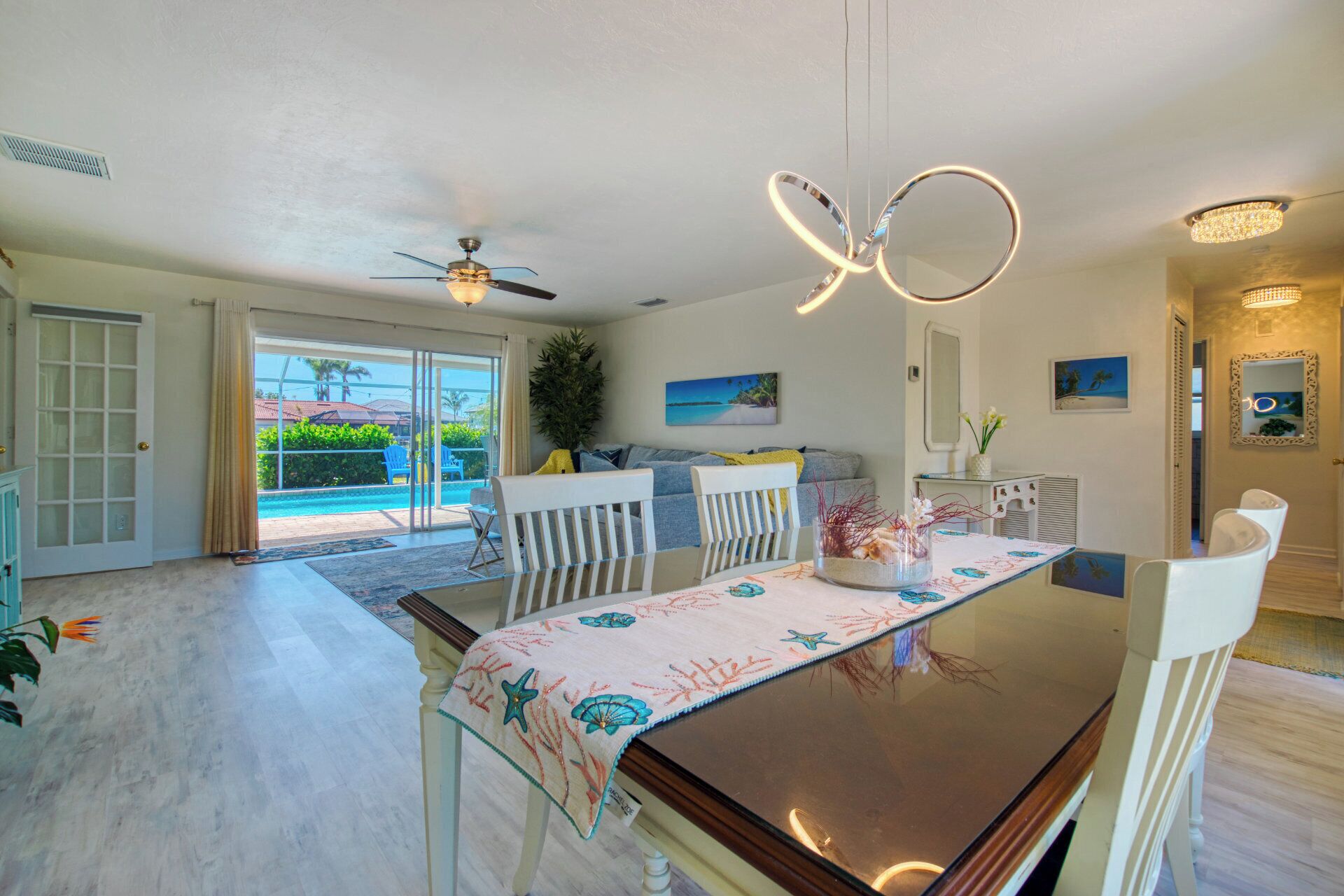 A dining area with a table set for two, a ceiling fan, and a view of a pool and greenery outside.