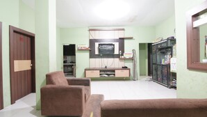 A living room with two brown sofas, a TV on a wooden cabinet, and a kitchen area in the background.