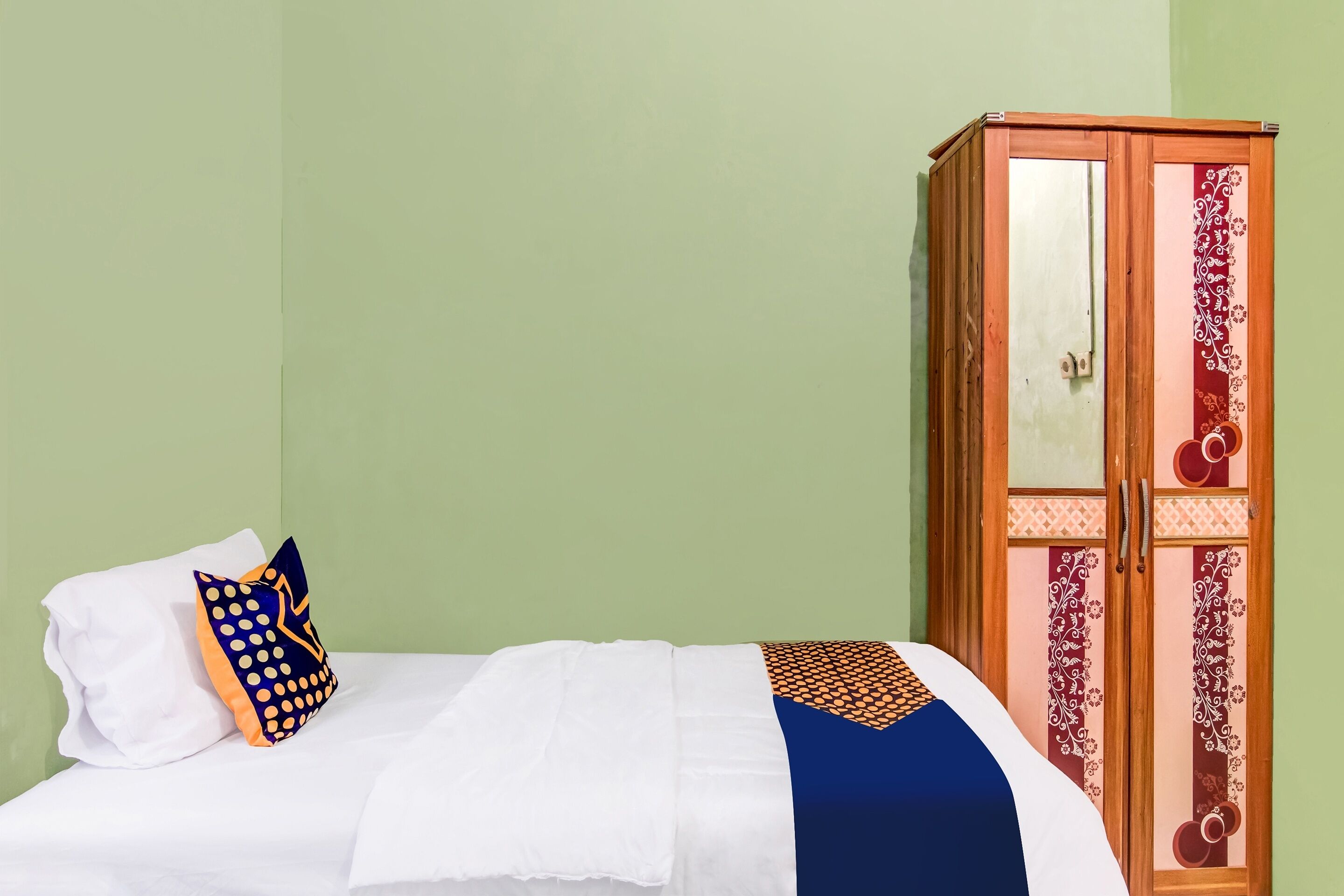 A neatly made bed with a white duvet and a decorative pillow, a wooden wardrobe with glass doors, and a green wall.