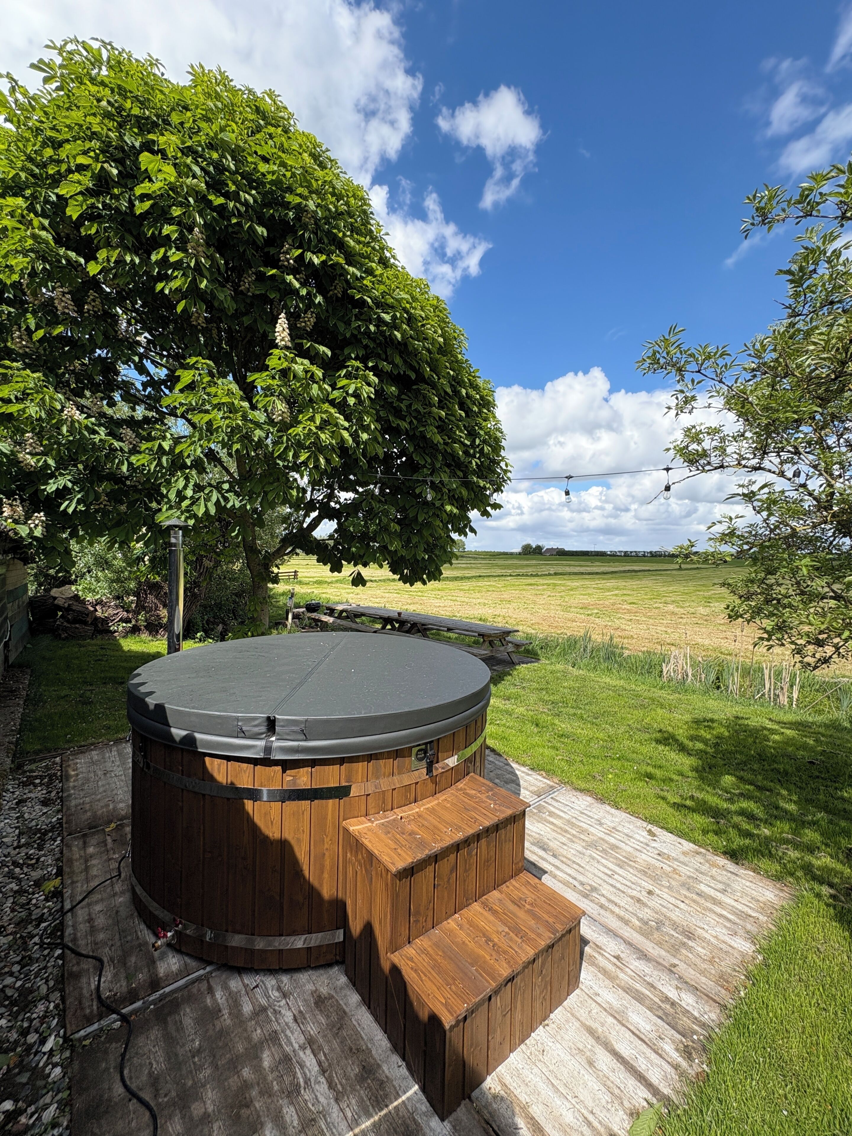 Un jacuzzi con revestimiento de madera, rodeado por una terraza también de madera, ubicado en un área con césped, un árbol grande y cielo despejado.