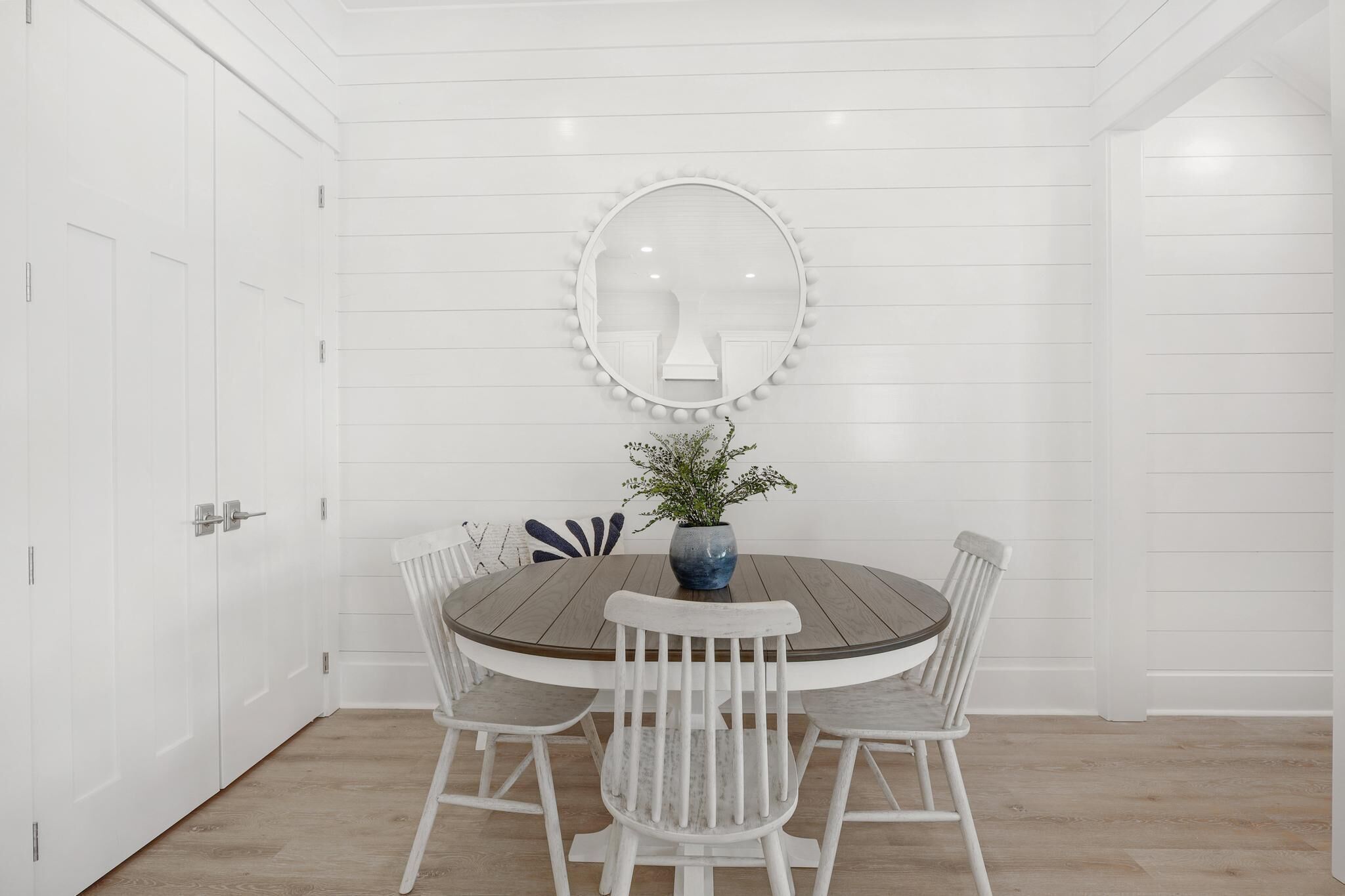 Une pièce blanche avec une table ronde en bois, des chaises blanches, un vase avec des fleurs et un miroir avec des lumières.