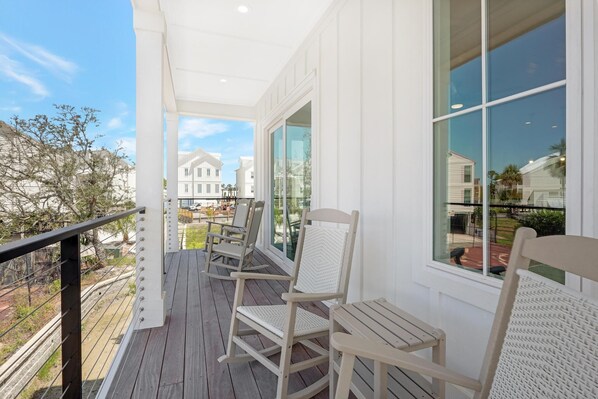 Relax in a serene outdoor space with a wooden deck, wicker rocking chairs, and a small table, surrounded by large windows offering views of the neighborhood.