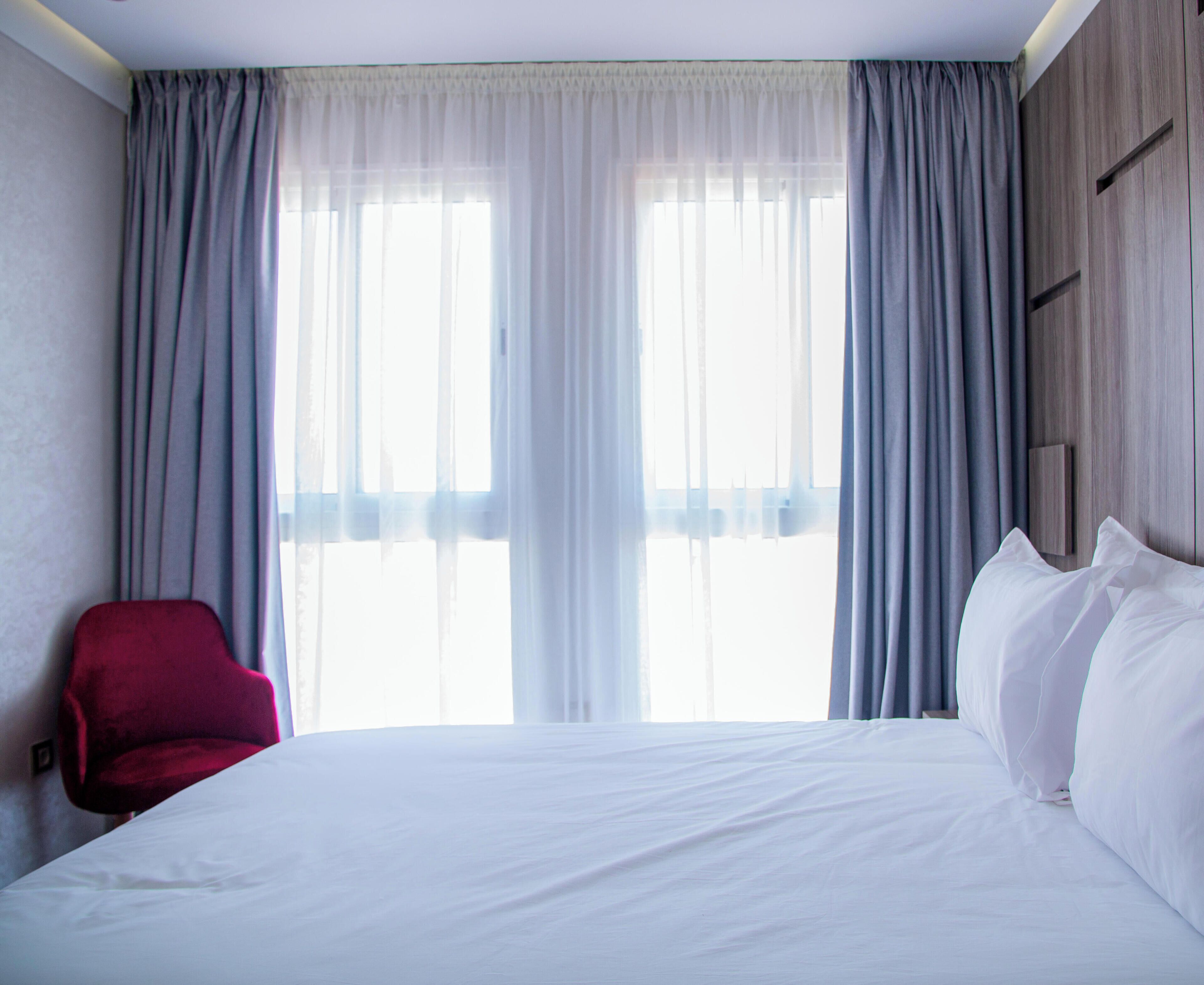 A hotel room with a bed, a red chair, and a large window with curtains.