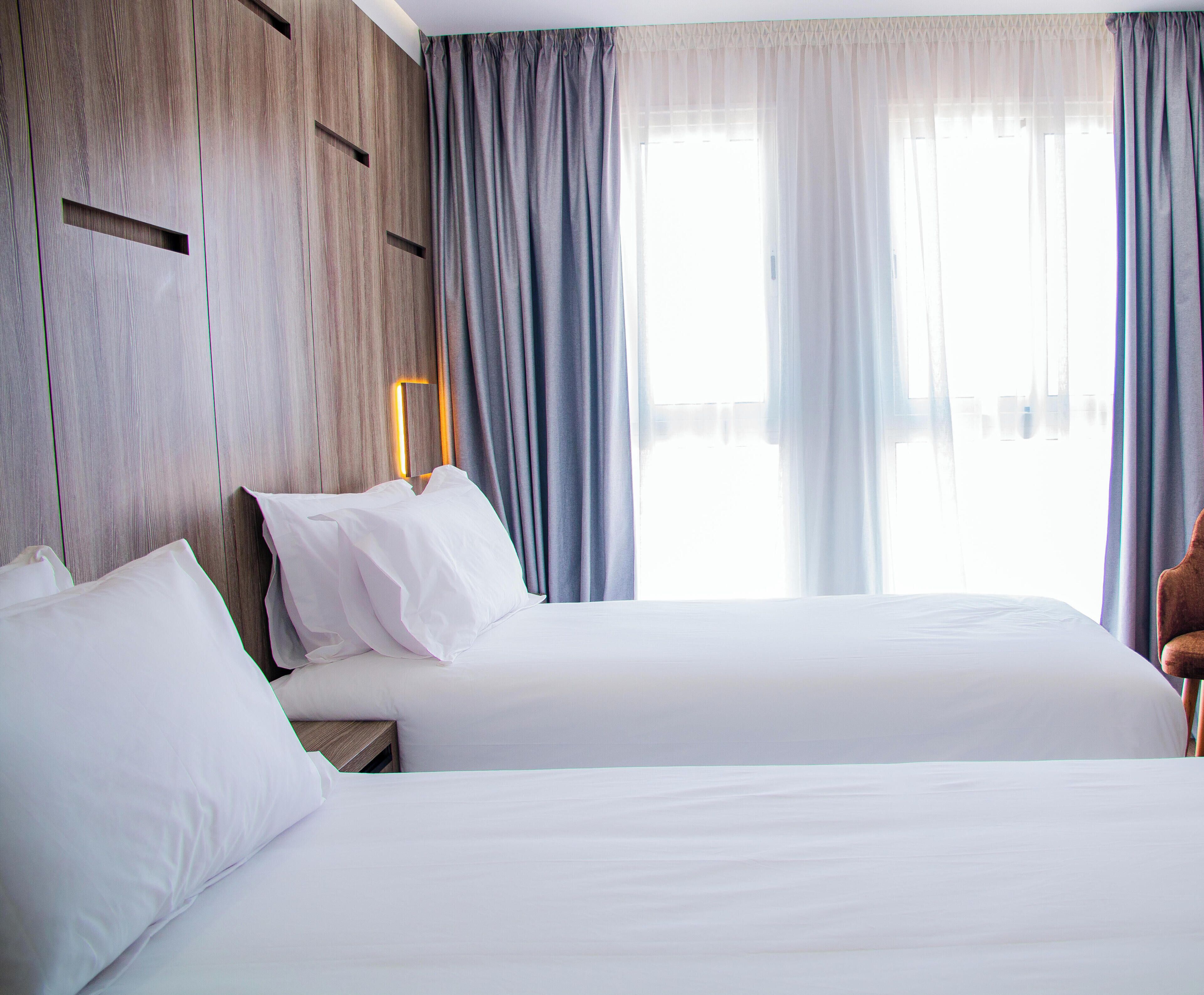 A hotel room with two beds, a wooden headboard, a nightstand, and a window with curtains.