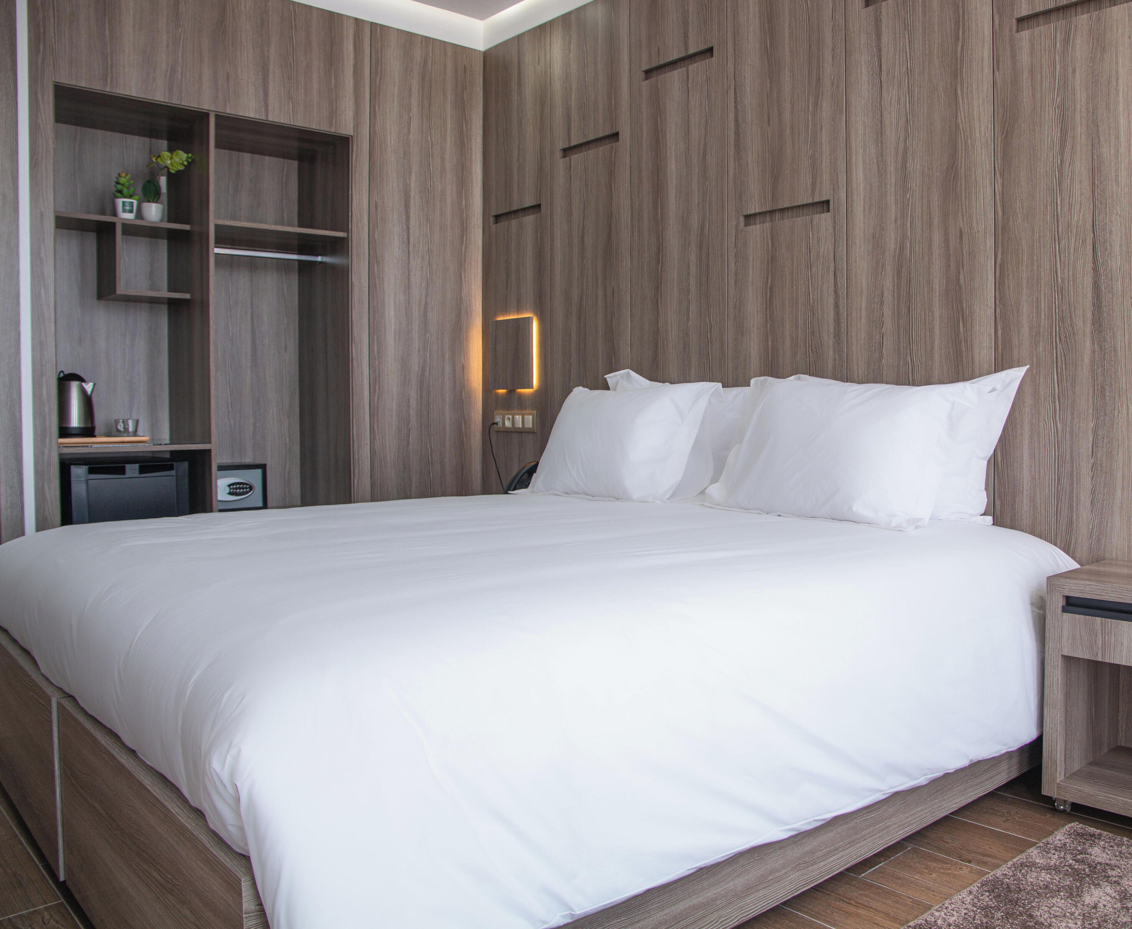 A modern hotel room with a large bed, wooden panel walls, built-in shelving, and a nightstand.
