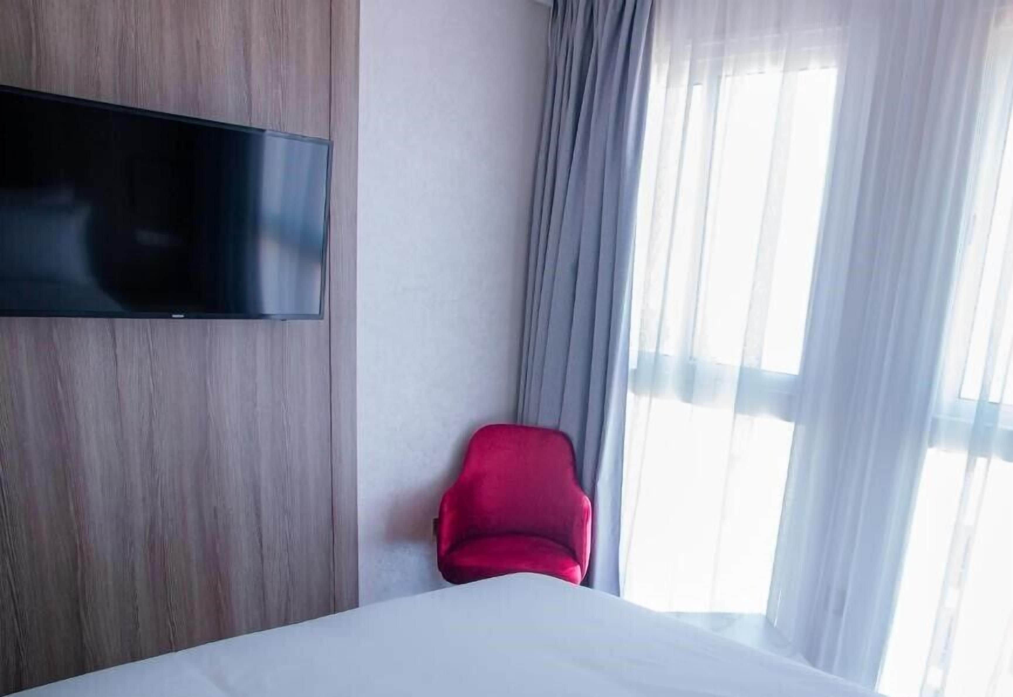 A hotel room with a bed, a red chair, a flat-screen TV mounted on the wall, and a window with sheer curtains.