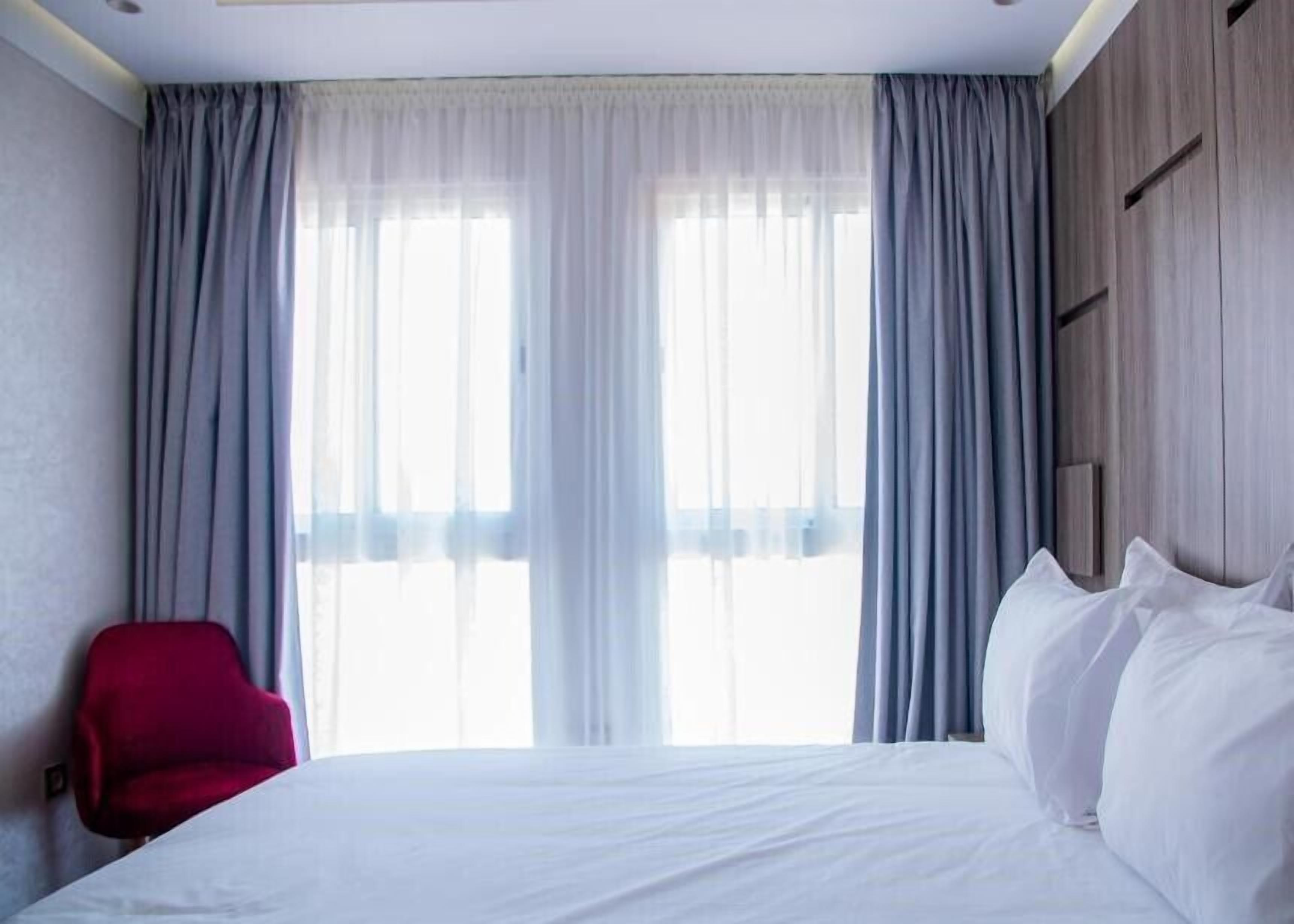 A hotel room with a bed, a red chair, and large windows with curtains.