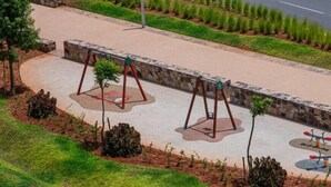 A playground with swings and a paved area, surrounded by grass and a stone wall. - Marchica Hotel (Nador)