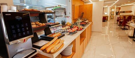 A breakfast buffet with a Nescafe coffee machine, various breads, pastries, and fruits.