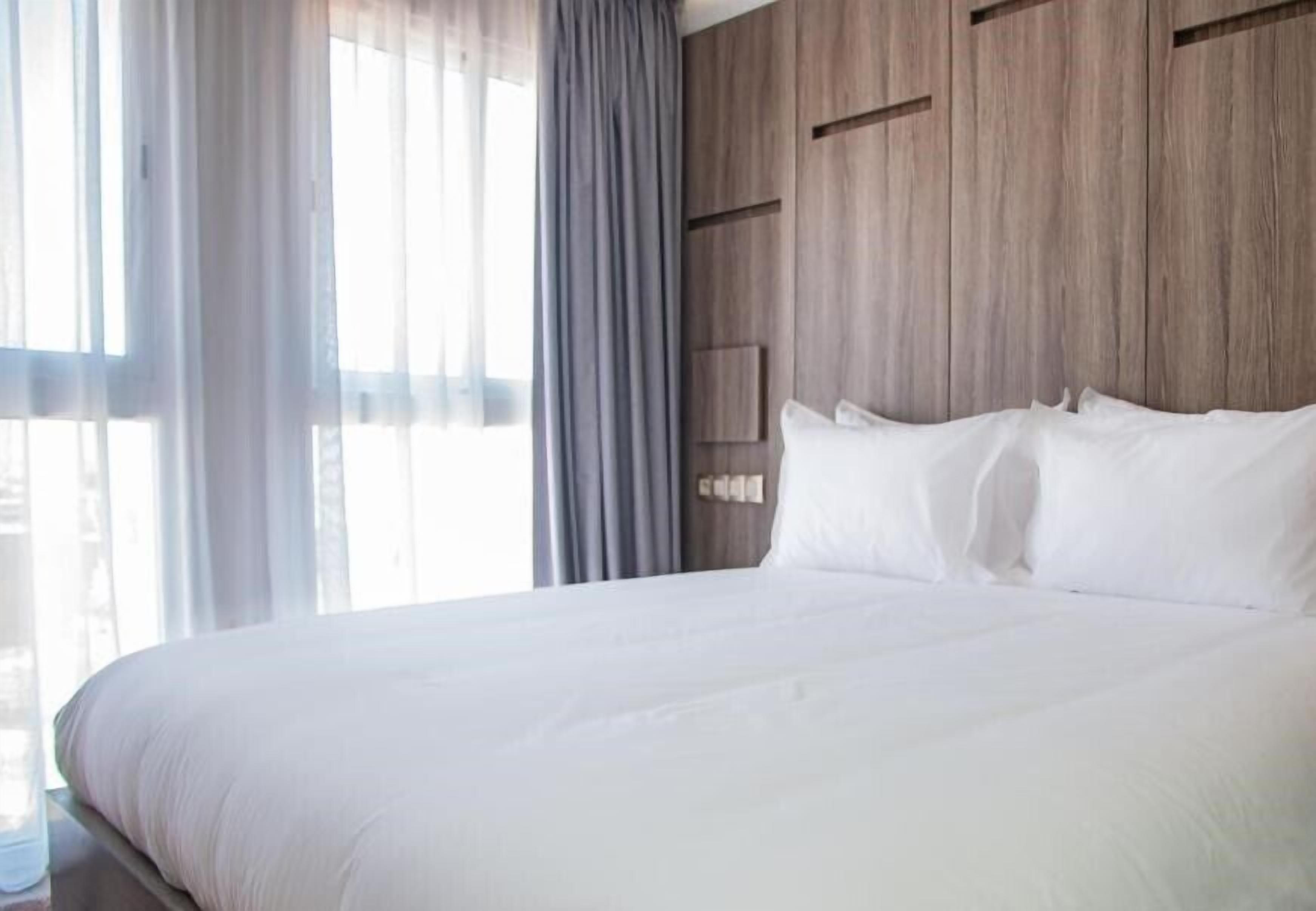 A large bed with white linens in a room with wooden paneling and a large window with sheer curtains.