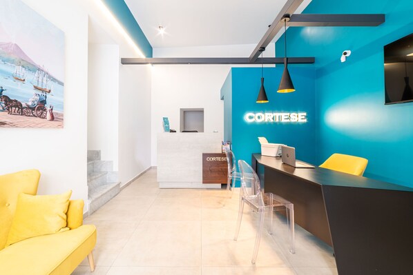 A modern reception area with a blue accent wall, a desk with a computer, and a yellow sofa. - Cortese Suite (Naples)
