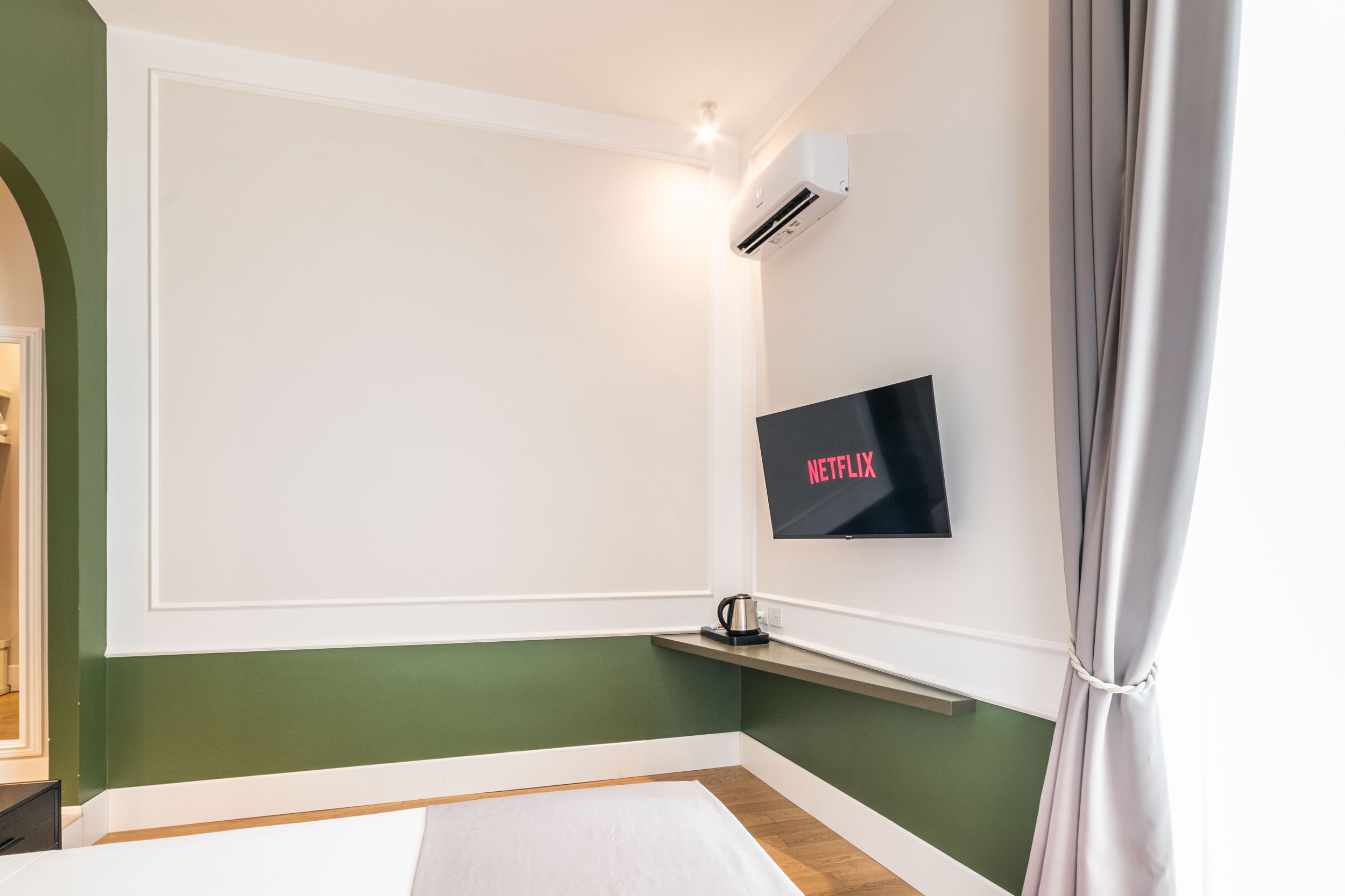 A hotel room with a bed, a mounted TV displaying 'NETFLIX', an air conditioning unit, and a small shelf.