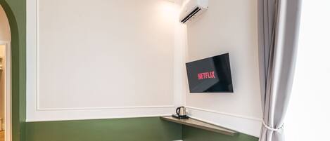 A hotel room with a bed, a mounted TV displaying 'NETFLIX', an air conditioning unit, and a small shelf.