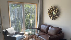 A modern living room with a brown leather sofa, a grey armchair with a furry throw, a glass coffee table, and a decorative wall piece. - Evo B&B - Adults Only, Waterfront (Northern Bruce Peninsula)