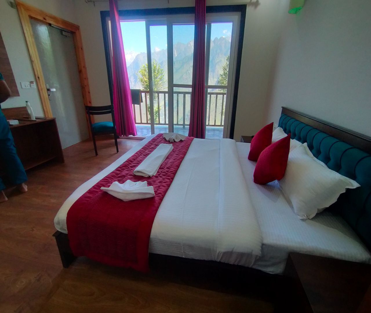 A hotel room with a large bed, a wooden door, a chair, a desk, and a view of mountains through the window.