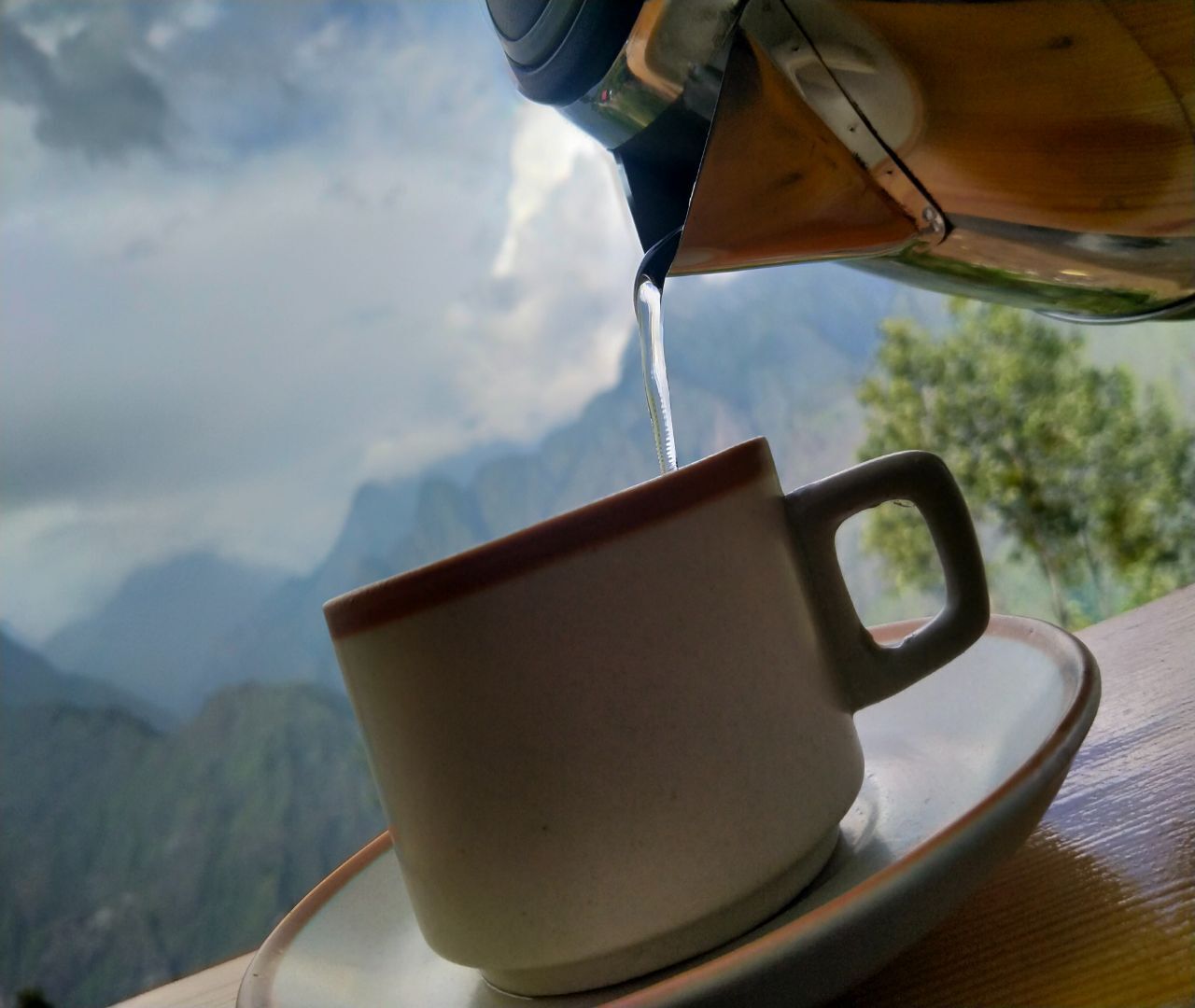 A cup of coffee being poured from a kettle, with a mountainous landscape in the background.