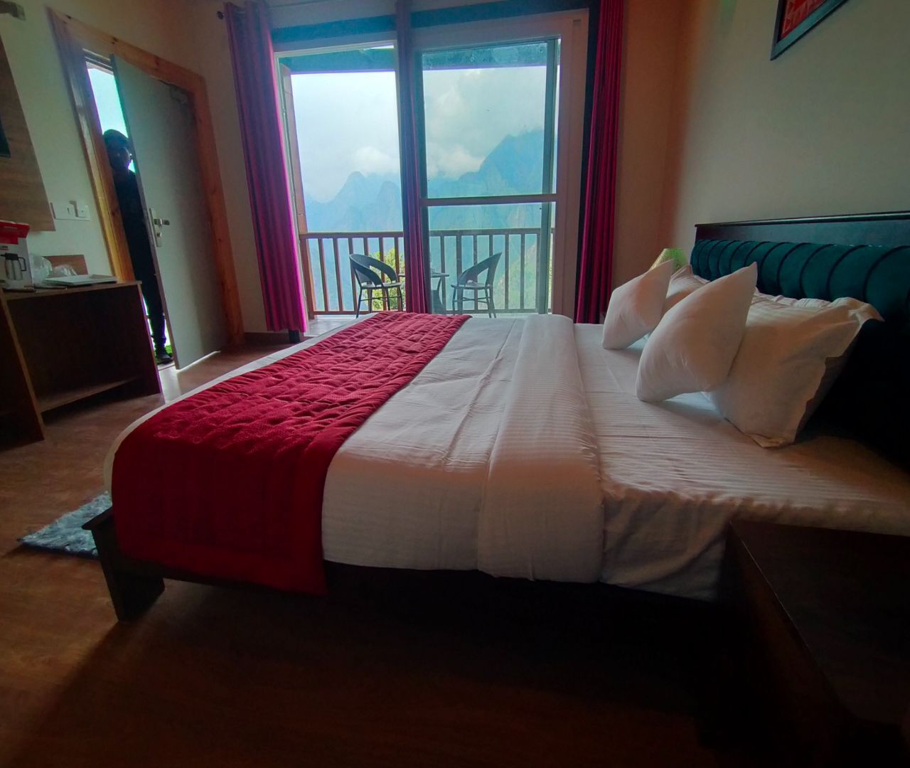 A bed with a red and white bedspread, a balcony with chairs, and a view of mountains.