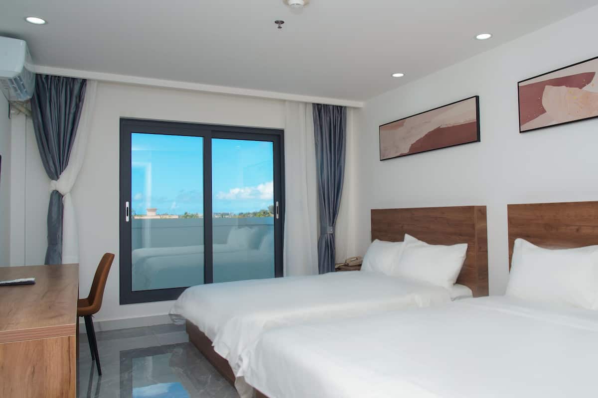 A hotel room with two beds, a desk, and a view of the sky through a glass door.