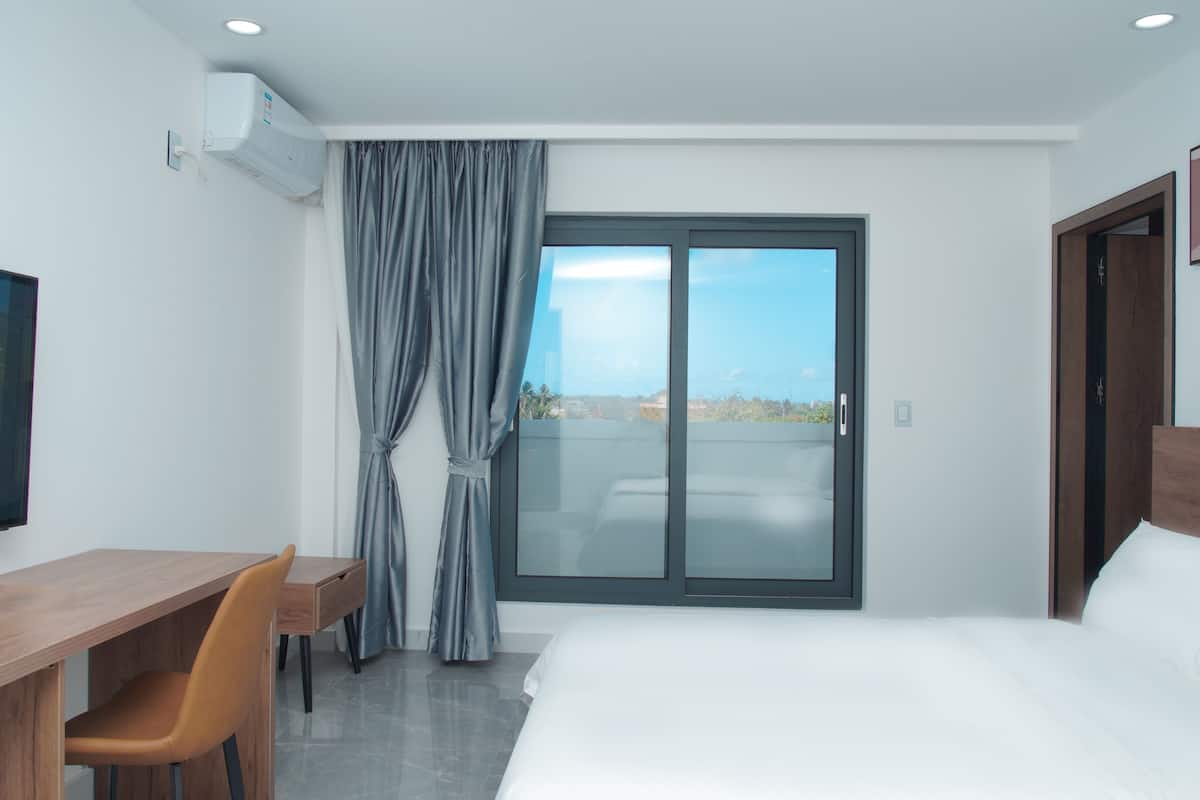 A hotel room with a bed, a desk with a chair, a TV, a window with curtains, and a door.