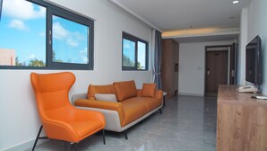 Una sala moderna con un sofá y una butaca, ventanas amplias, un escritorio de madera con un teléfono, y una puerta al fondo.