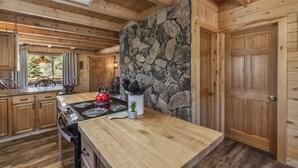 A wooden cabin kitchen with a stone wall, wooden cabinets, a stove, and a kettle on the counter. - Three-Story Home -1/8 of a mile of Fishing the Rio Grande River (Creede)