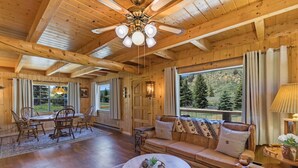 A cozy cabin interior with a wooden ceiling, a dining area with chairs and a table, a sofa with cushions, and a large window offering a view of trees. - Three-Story Home -1/8 of a mile of Fishing the Rio Grande River (Creede)