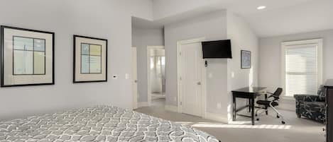 A bedroom with a large bed, a desk with a chair, a TV mounted on the wall, and a door leading to another room.