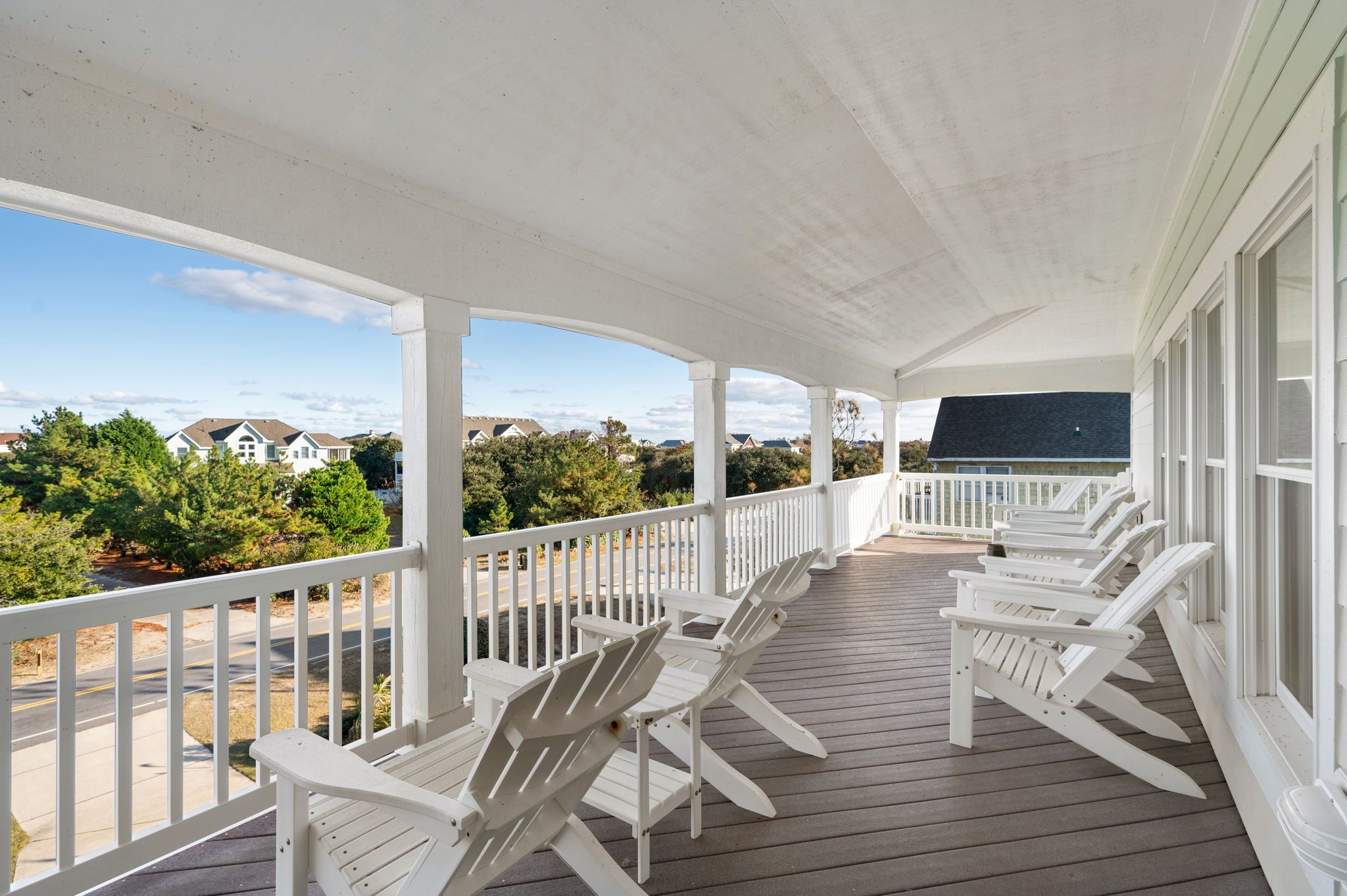 A covered porch with white railings and Adirondack chairs, overlooking a residential area with houses and trees.