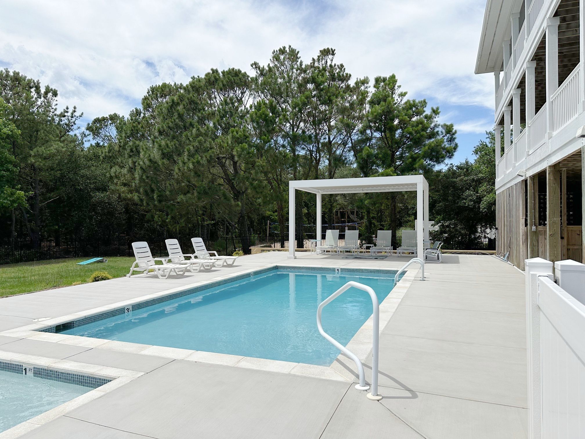 A rectangular swimming pool with a white deck, surrounded by lounge chairs and a covered seating area.