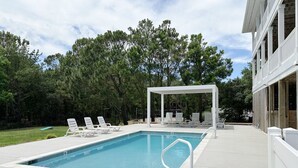 A rectangular swimming pool with a white deck, surrounded by lounge chairs and a covered seating area.