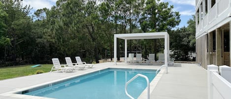 A rectangular swimming pool with a white deck, surrounded by lounge chairs and a covered seating area.