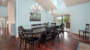 A spacious dining area with a large round table and multiple chairs, a chandelier, and a painting on the wall.