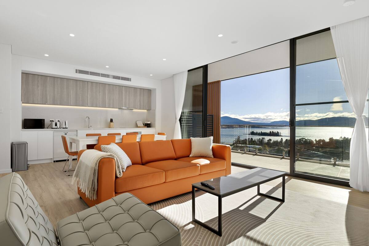 A modern living room with a large window, a sofa, a coffee table, and a kitchen area.
