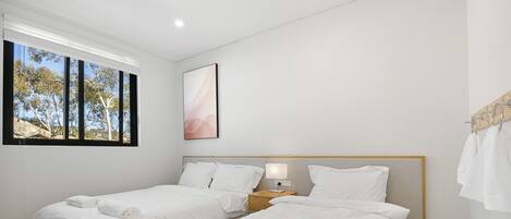 A hotel room with two beds, a wooden headboard, a bedside table with a lamp, and a window with a view of trees.