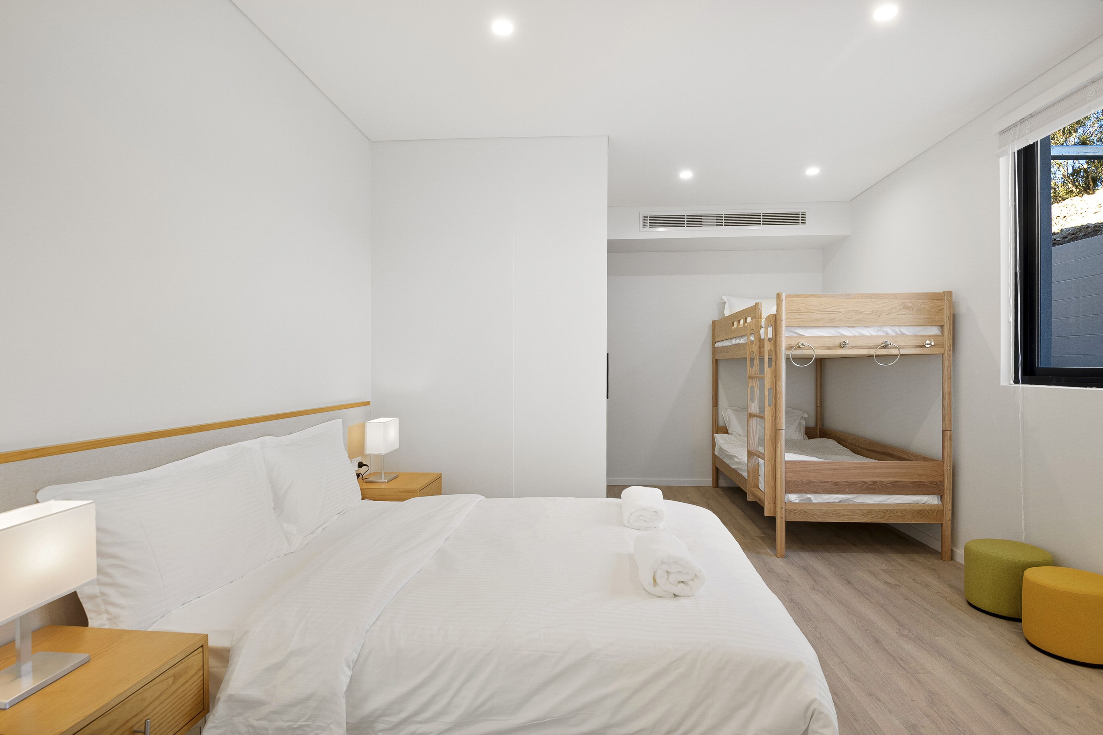 Moderni hotellihuone, jossa on suuri sänky, puinen kerrossänky, yöpöytä lampulla ja ikkuna, josta on näkymä.