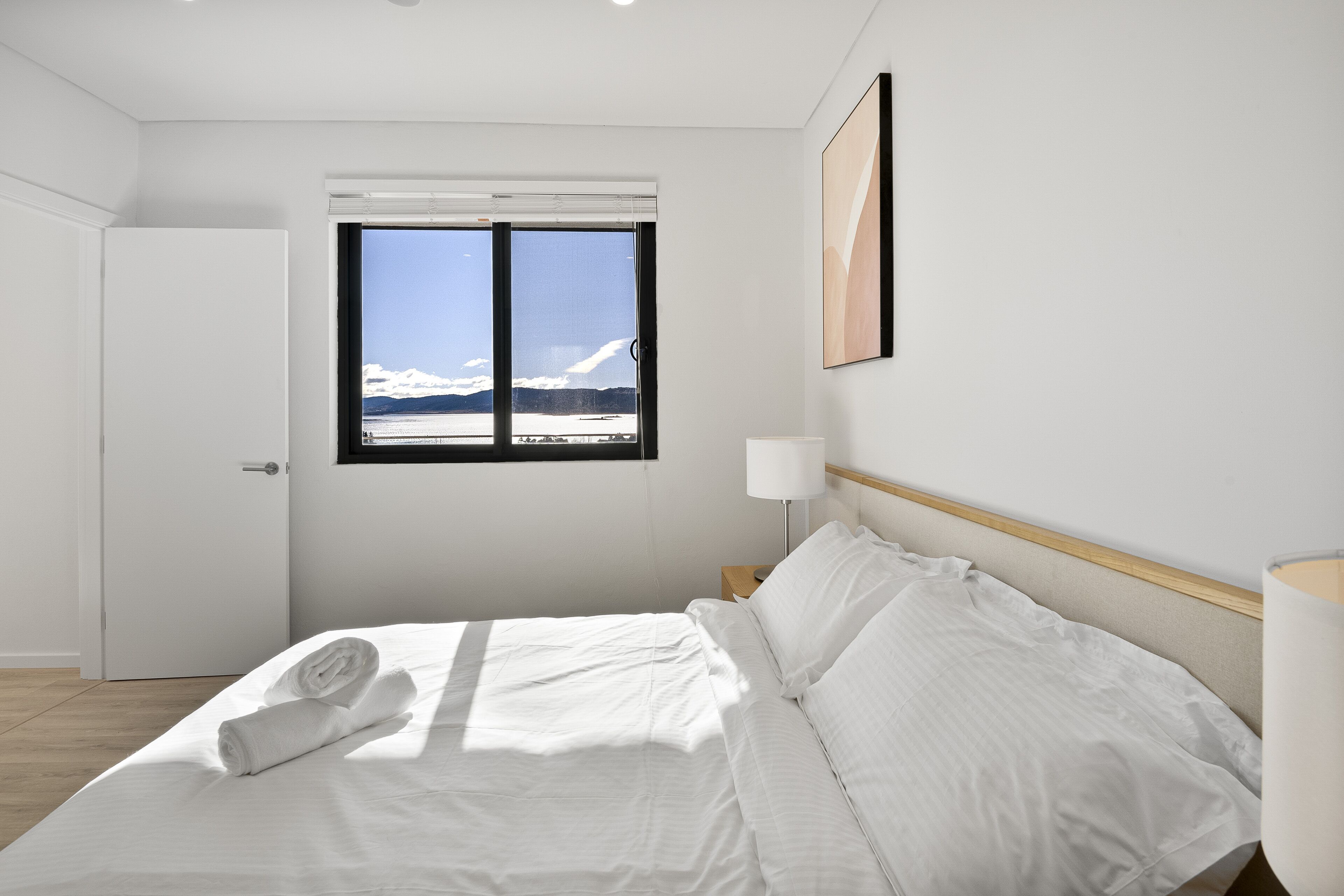 A hotel room with a bed, a window with a view of mountains, a door, and a nightstand with a lamp.