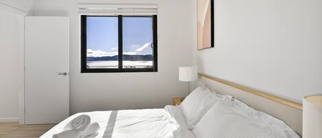 A hotel room with a bed, a window with a view of mountains, a door, and a nightstand with a lamp.