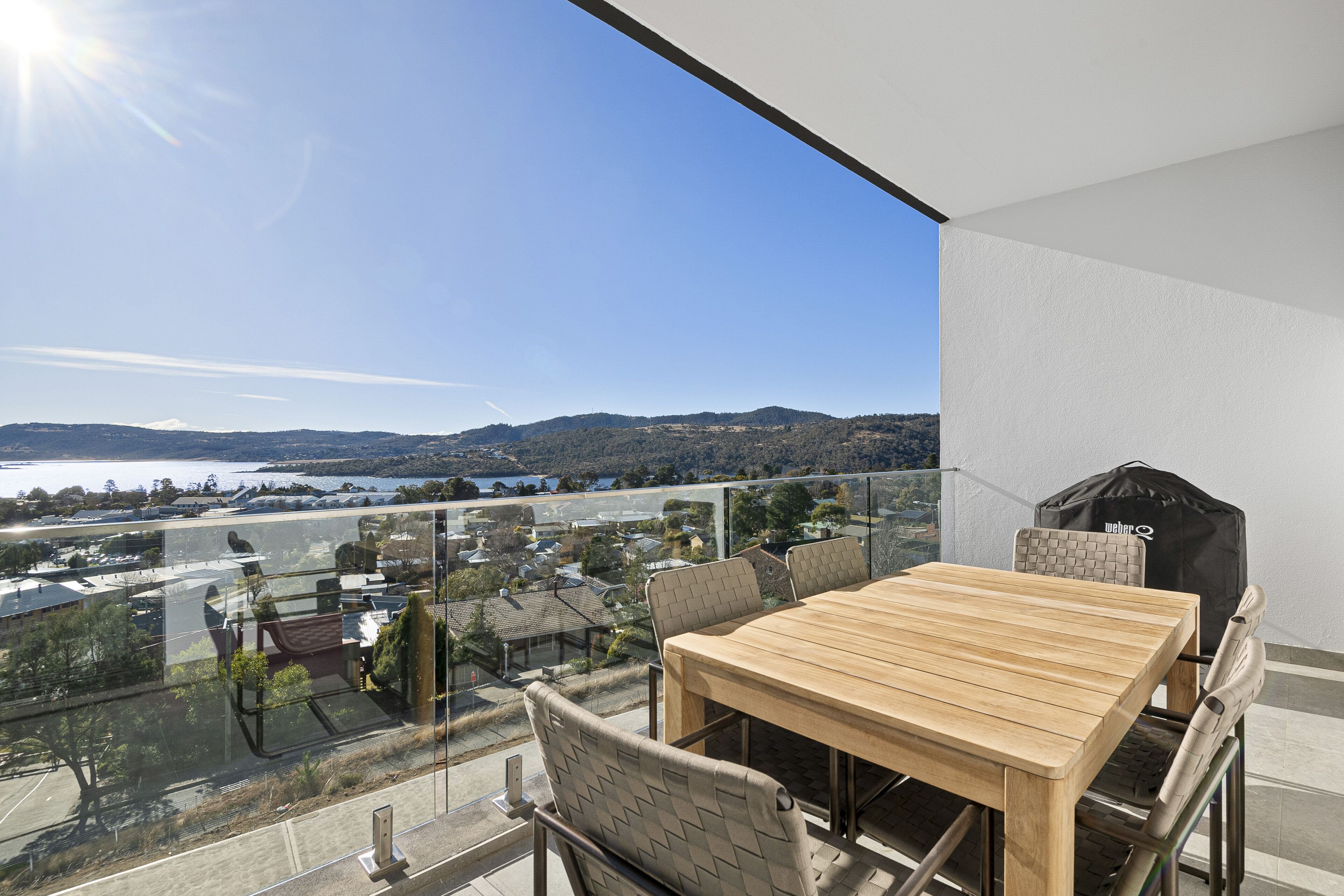 A balcony with a wooden table and chairs, overlooking a scenic view of a lake and distant hills.