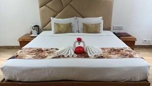 A neatly made bed with a decorative swan pillow arrangement, a wooden nightstand with a coffee maker, and a bedside table with a lamp and a book. - Hotel Grand Riviera (Paonta Sahib)