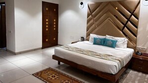 A modern bedroom with a large bed, a headboard with a geometric pattern, a wooden wardrobe, and a patterned rug. - Hotel Grand Riviera (Paonta Sahib)