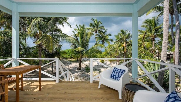 Un espacio al aire libre techado con cubierta de madera, mobiliario blanco y vista a palmeras y la playa.