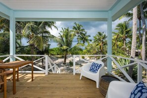 A covered outdoor area with a wooden deck, white furniture, and a view of palm trees and beach. - Brand New! Private Beachfront Cottage on World-Famous French Leave Beach (Governors Harbour)