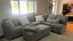Un canapé modulable assorti d’un pouf, une grande fenêtre donnant sur la verdure et un porte-manteau avec des vêtements et des chaussures.