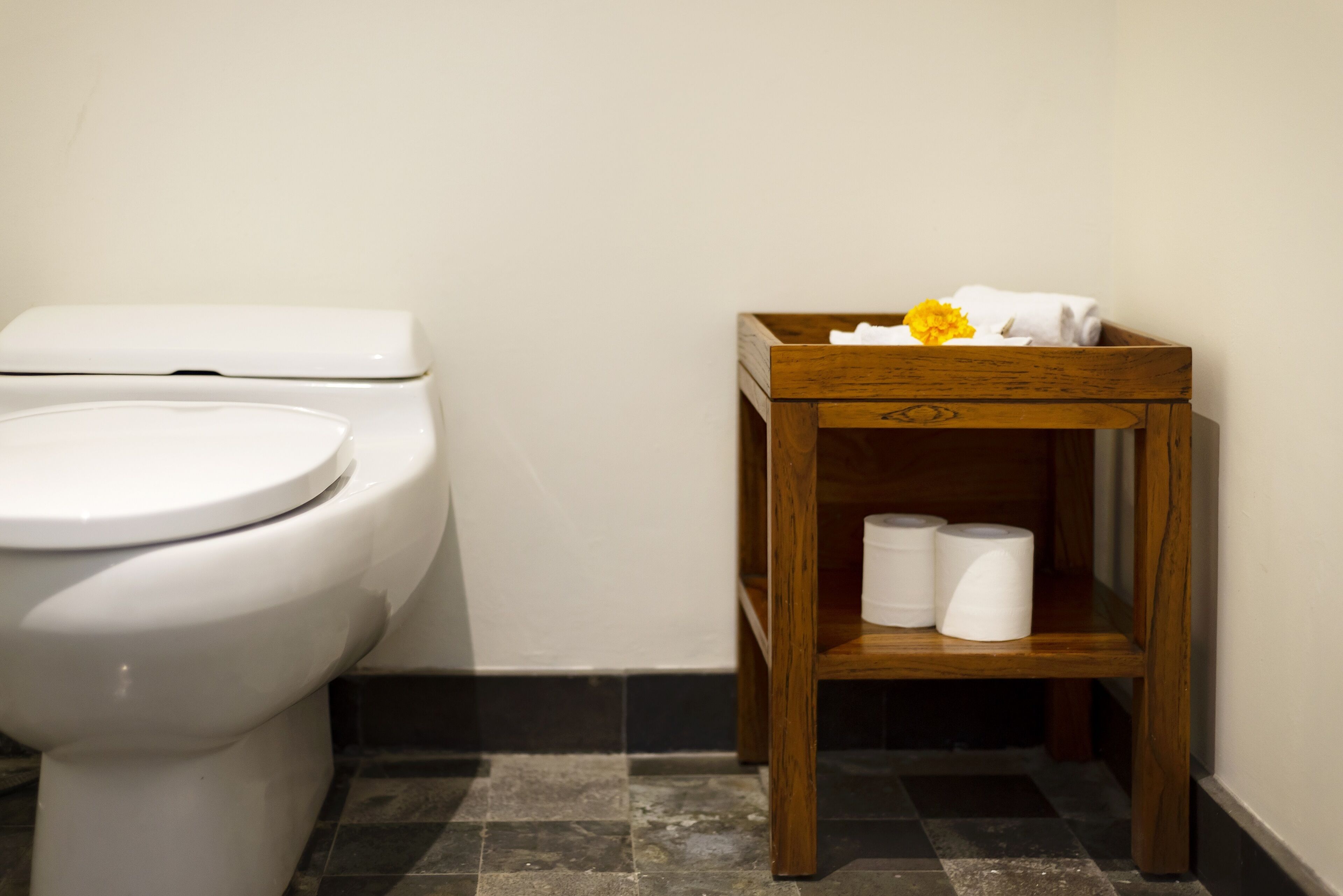 Un baño con inodoro blanco, un estante de madera con toallas y papel higiénico, y suelo de baldosas.