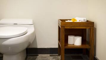 Un baño con inodoro blanco, un estante de madera con toallas y papel higiénico, y suelo de baldosas.