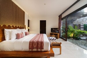 A bedroom with a large bed, a wooden bench, a TV, and a view of a garden through large windows.