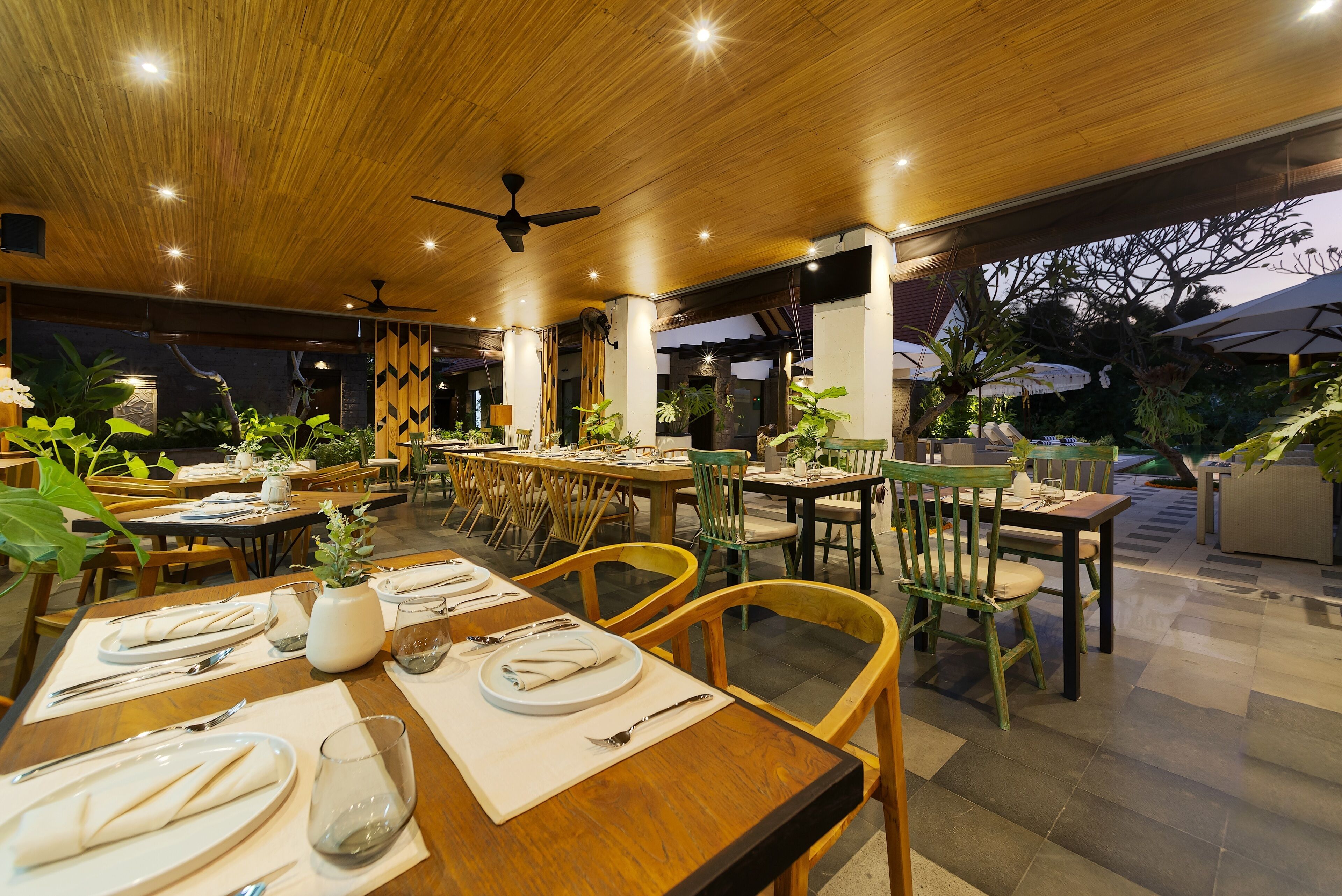 Un restaurante con mesas y sillas de madera, ventiladores de techo y plantas en macetas.