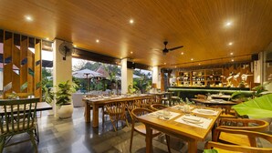 An outdoor dining area with wooden tables and chairs, potted plants, and a bar area with shelves of bottles.
