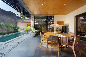 Uma área de refeições moderna ao ar livre, com piscina, móveis de madeira e plantas em vasos.